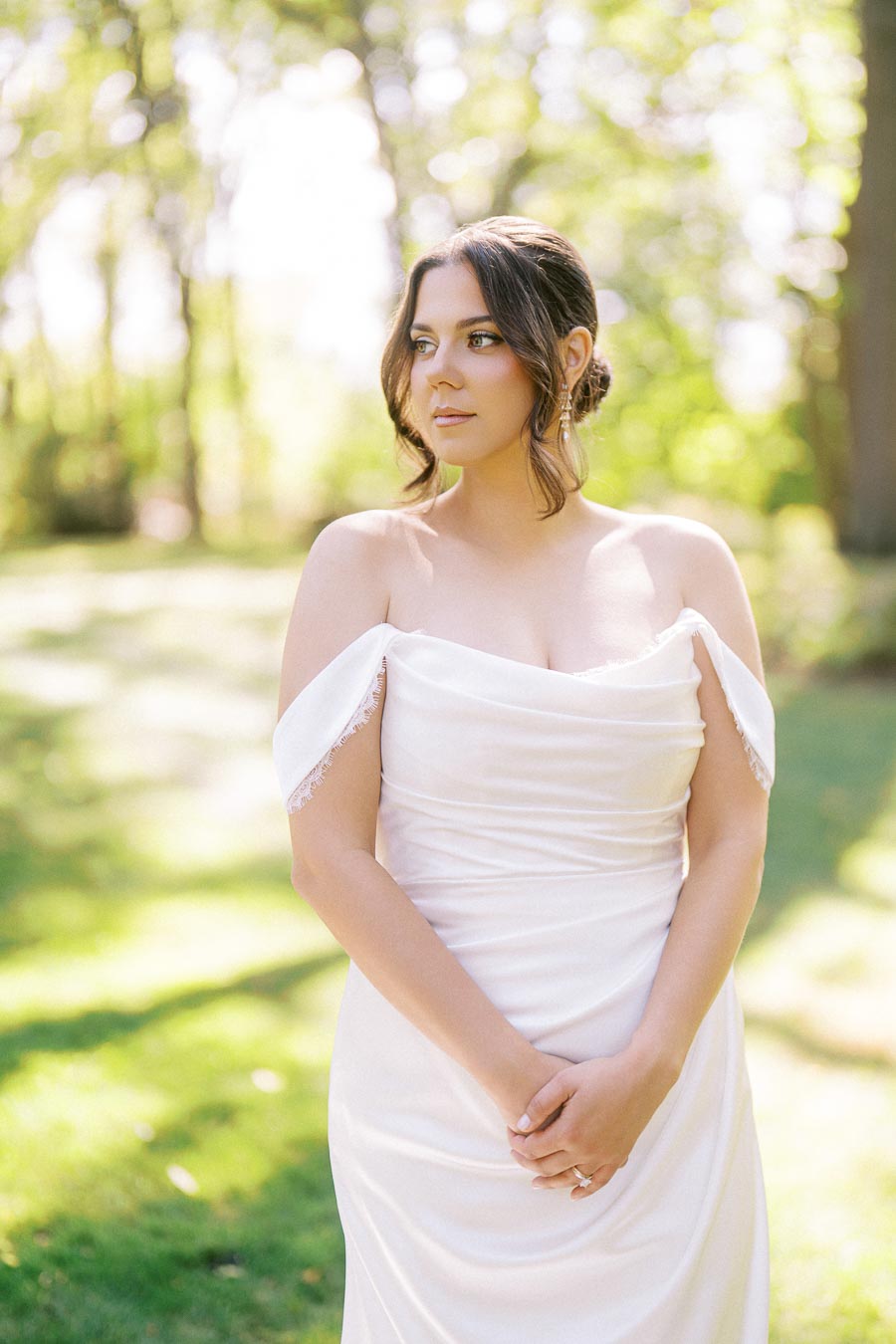 A bride in an elegant off-shoulder white wedding dress stands outdoors in a sunlit garden, surrounded by lush, green trees, looking serene and thoughtful.