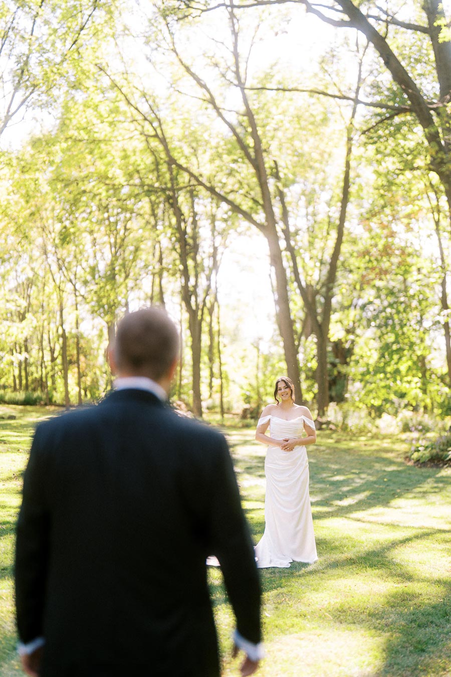 A bride in a white dress standing on a sunlit grassy lawn with trees, facing a groom in a black suit for a first look at their outdoor wedding.