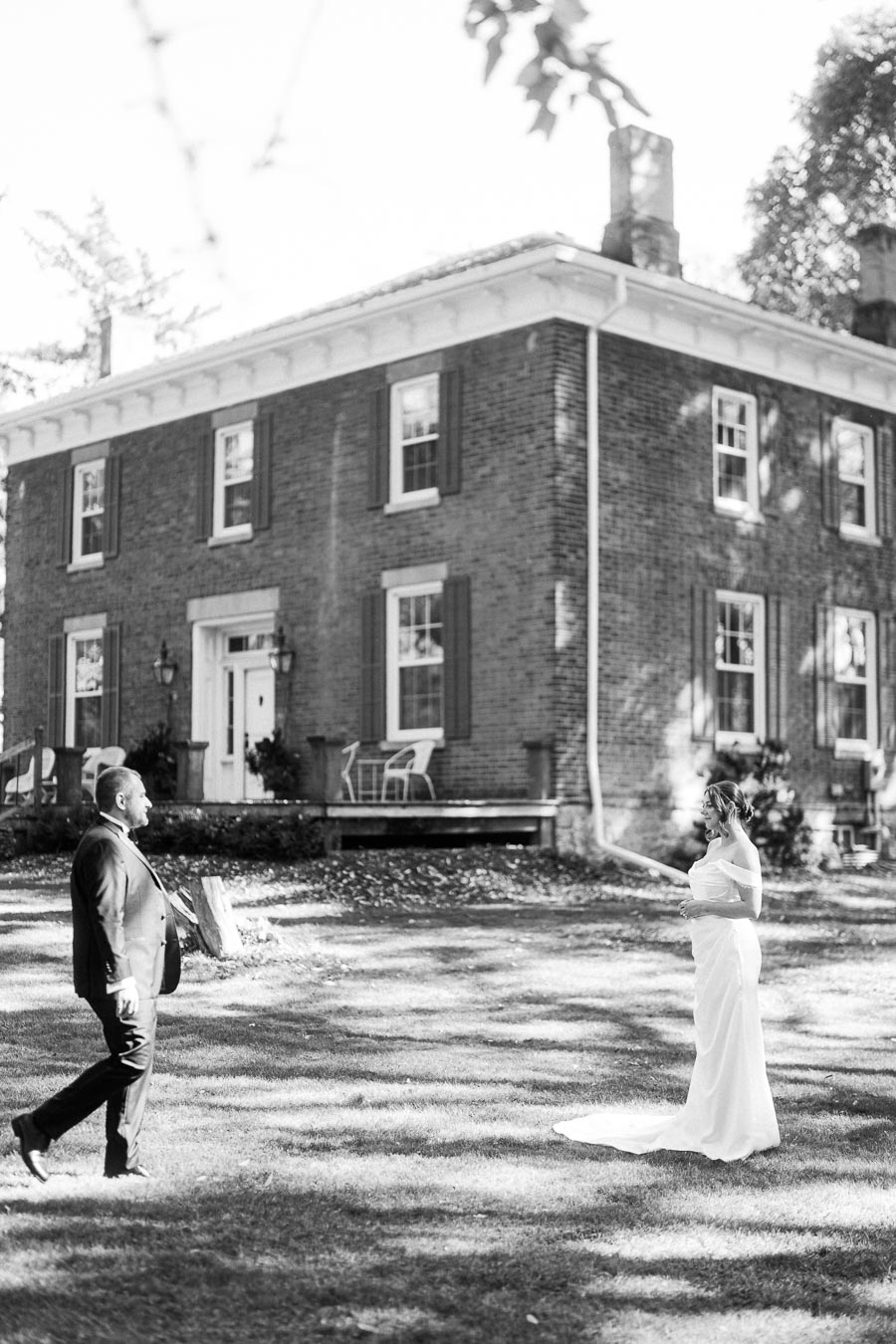 Black and white photo of a bride in a white gown and groom in a suit, sharing a moment on the lawn of a historic brick building, emphasizing romance and timeless wedding memories.