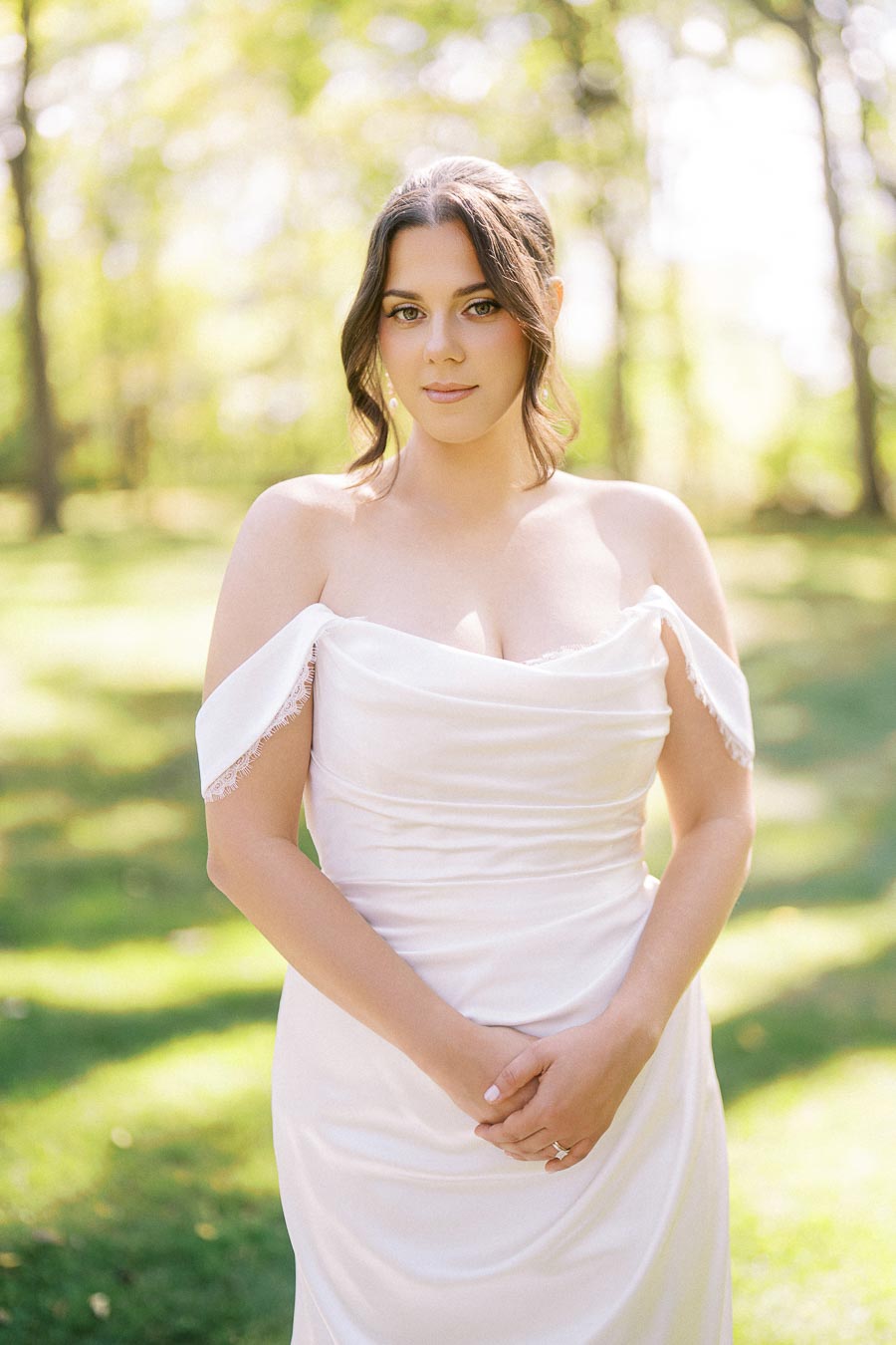 A bride in an elegant off-the-shoulder white wedding dress standing outdoors with a blurred green forest background, under soft natural sunlight.