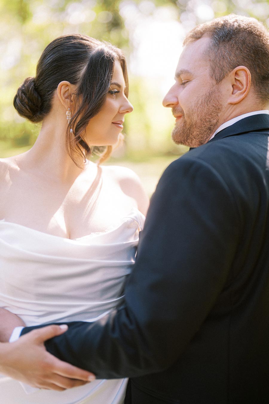 Couple Embracing on Wedding Day in Outdoor Setting with Bride in Elegant White Gown and Groom in Black Suit, Captured in Natural Light