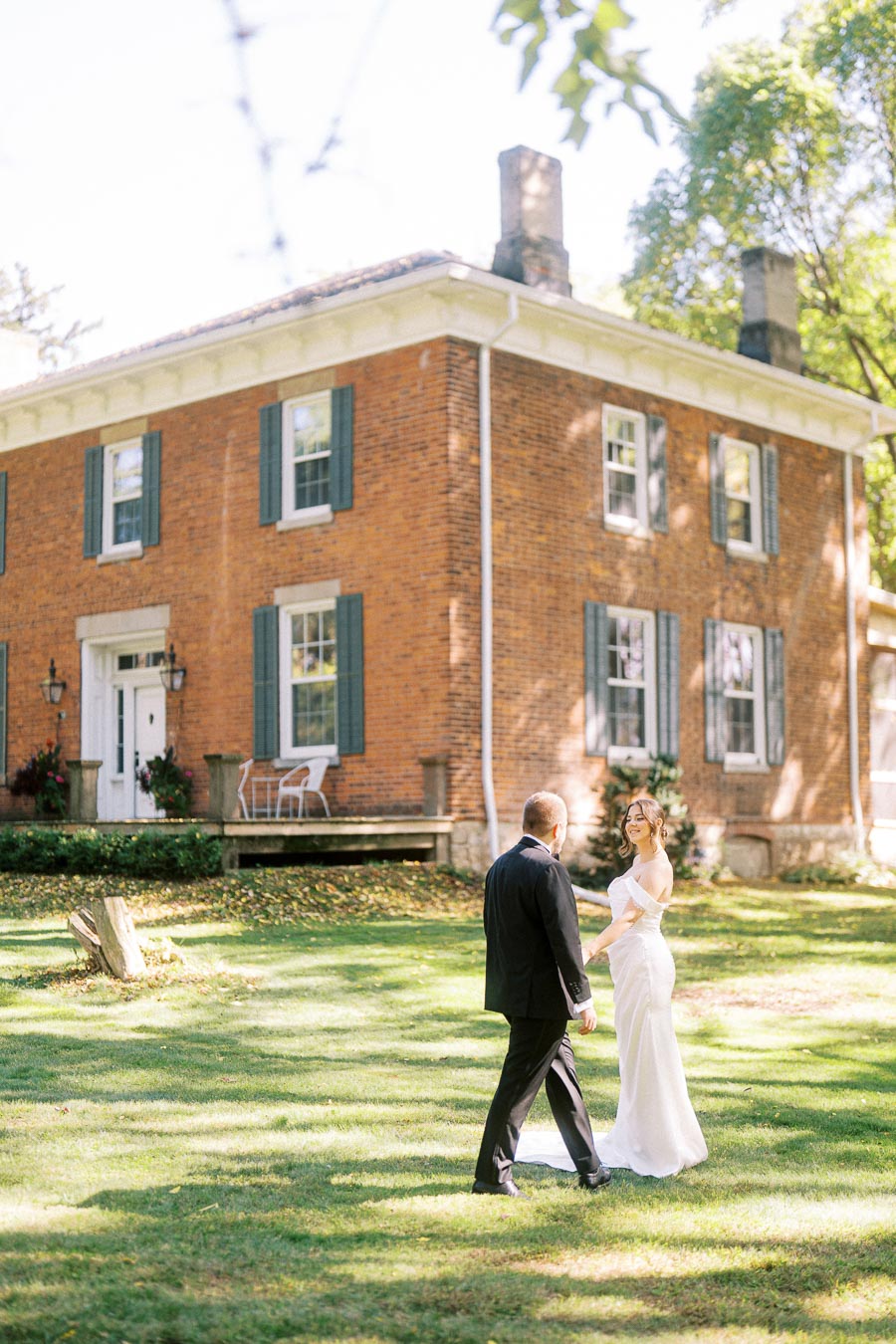 Elegant bride and groom in a white dress and black suit holding hands in front of a charming brick house with blue shutters and lush greenery, capturing a picturesque outdoor wedding moment.