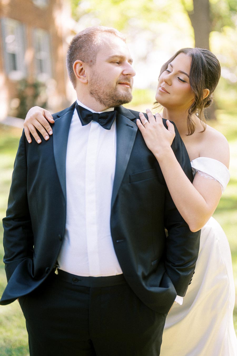 A bride embracing a groom from behind, both in formal wedding attire, standing in a sunlit garden setting.