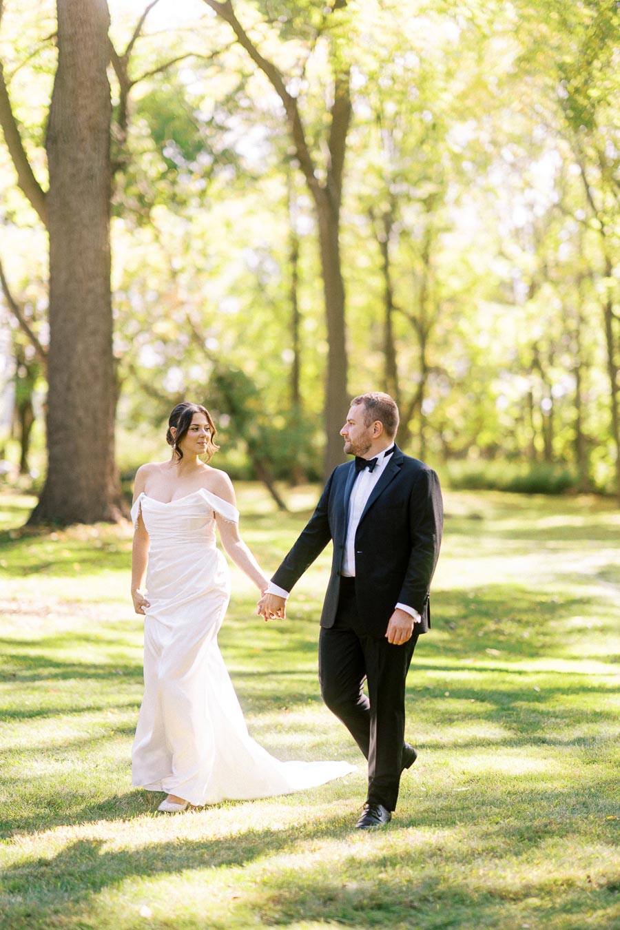 A bride in a white wedding dress walks hand in hand with a groom in a black suit through a sunny forested park, capturing a romantic moment on their special day.