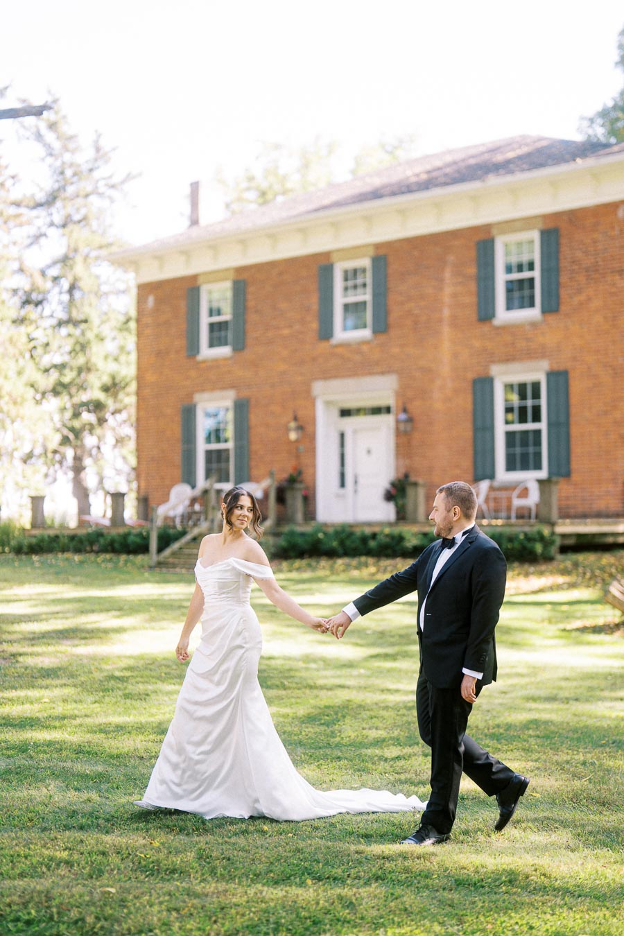 A bride in a white gown and a groom in a black tuxedo holding hands and walking on a grassy lawn, in front of a charming brick house with green shutters, on a sunny day.