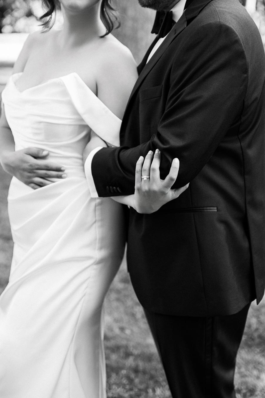 Black and white photo of a couple in elegant wedding attire, with the bride in a strapless gown and the groom in a suit, embracing outdoors.