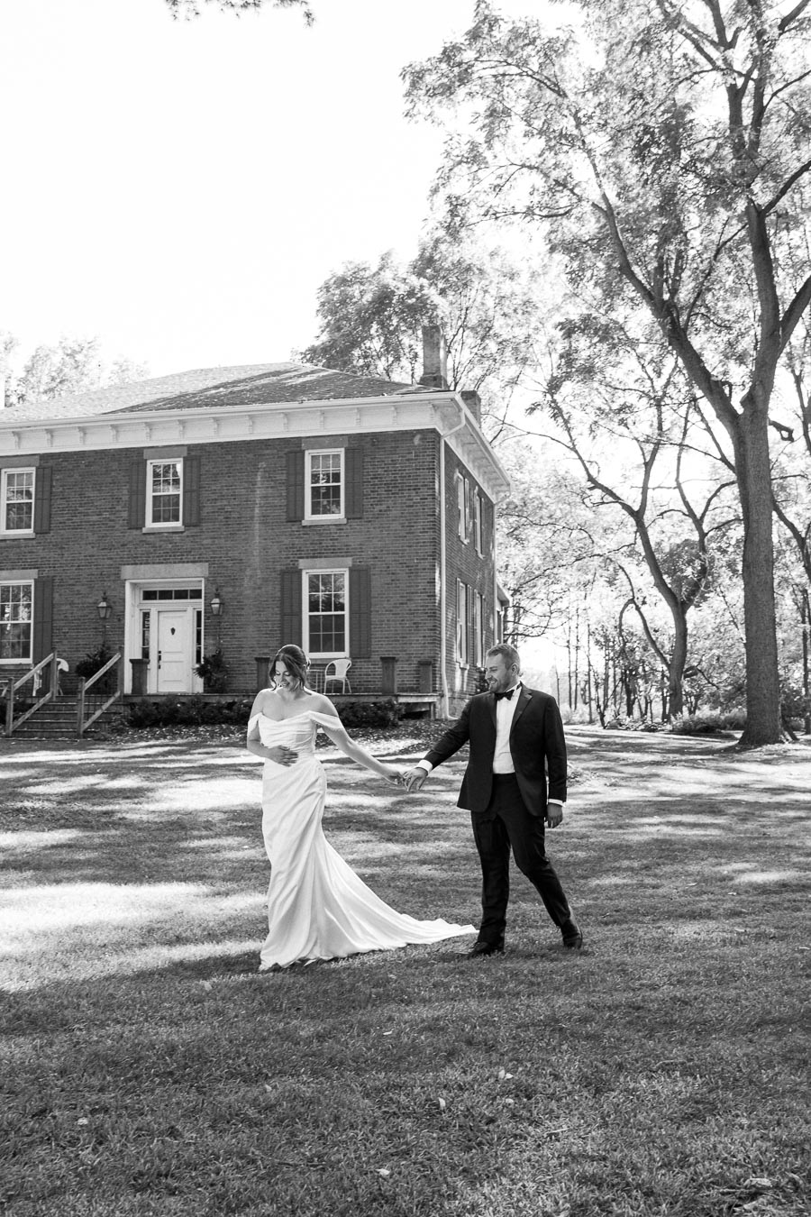 Black and white photo of a couple holding hands, wearing wedding attire in front of a historic brick house with large trees and a grassy lawn, capturing an elegant moment of joy and connection on their special day.