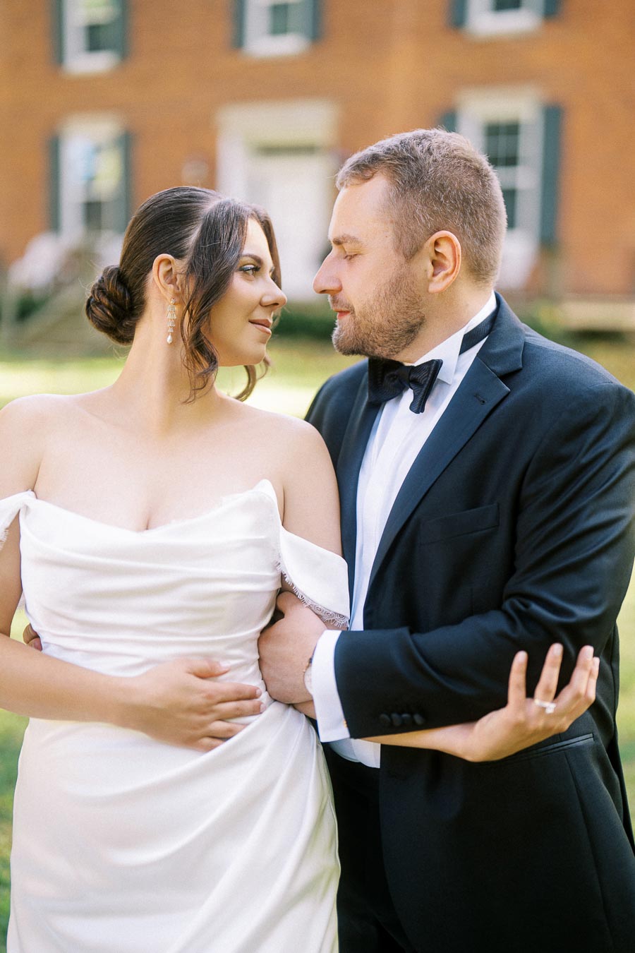 A bride in a white dress and groom in a black suit lovingly embrace in front of a brick building, showcasing a romantic wedding day moment.