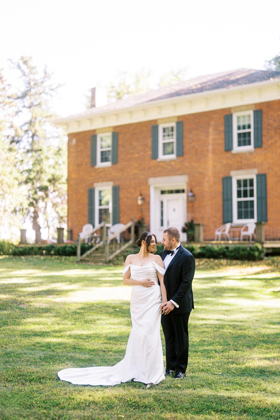 Elegant married couple standing in front of a charming brick house on a sunny day, with the bride in a flowing white gown and the groom in a classic black tuxedo, capturing a romantic moment on their wedding day.