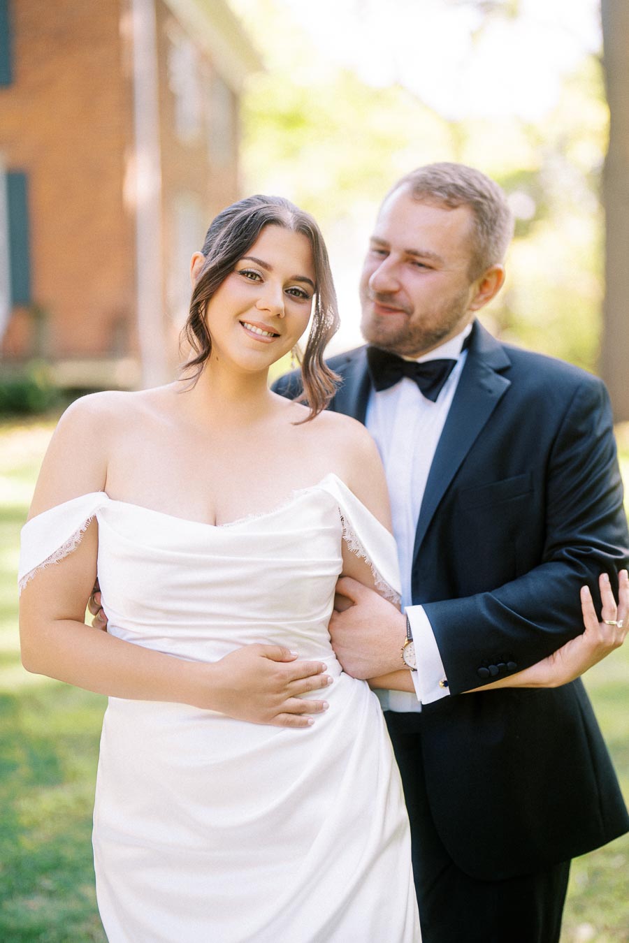 A bride in an elegant off-shoulder wedding dress smiles while being embraced by a groom in a classic black tuxedo in a sunny outdoor setting with greenery and a brick building in the background.