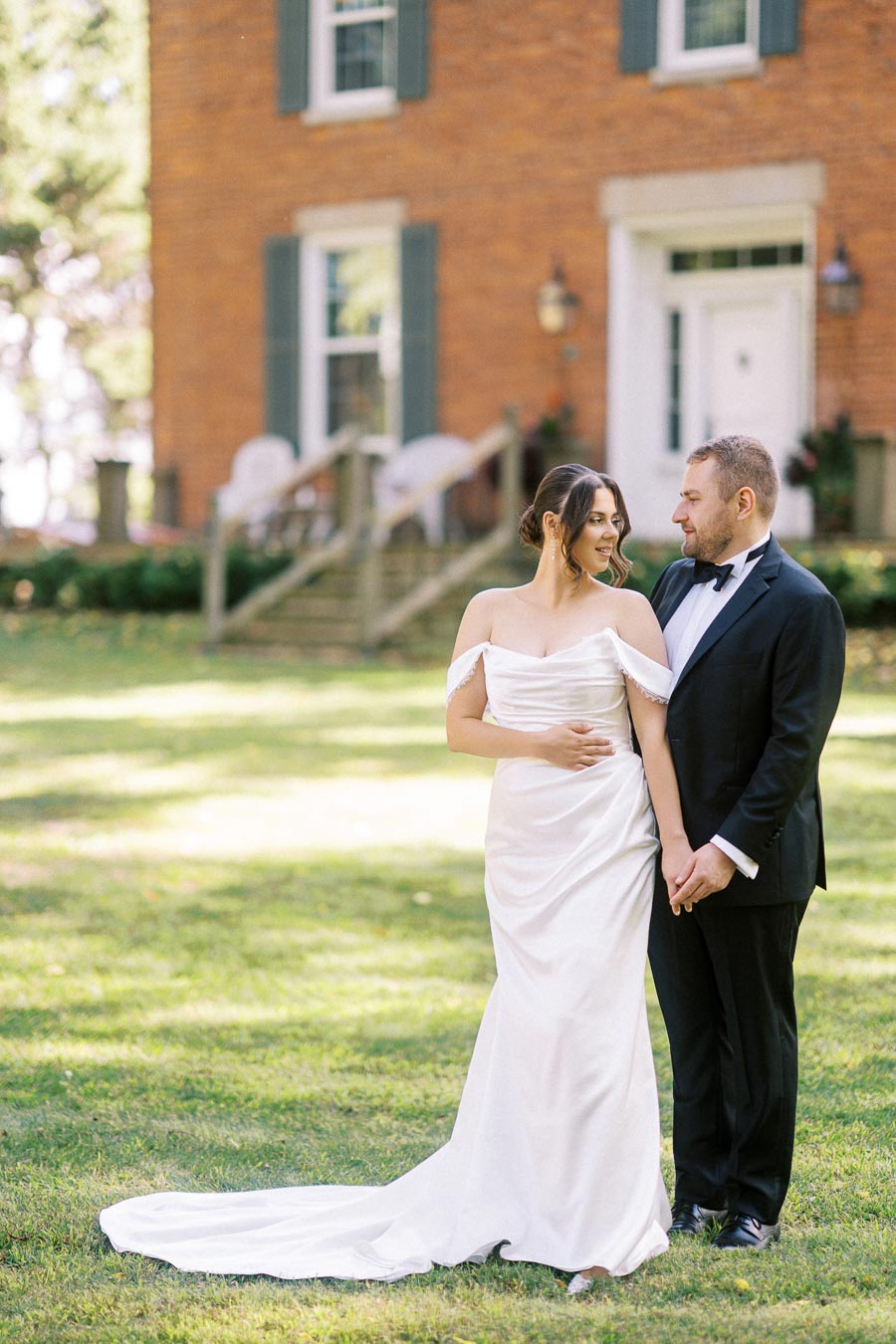 Elegant couple standing together in front of a historic brick building with greenery, bride in a white dress and groom in a black tuxedo, outdoor wedding setting.