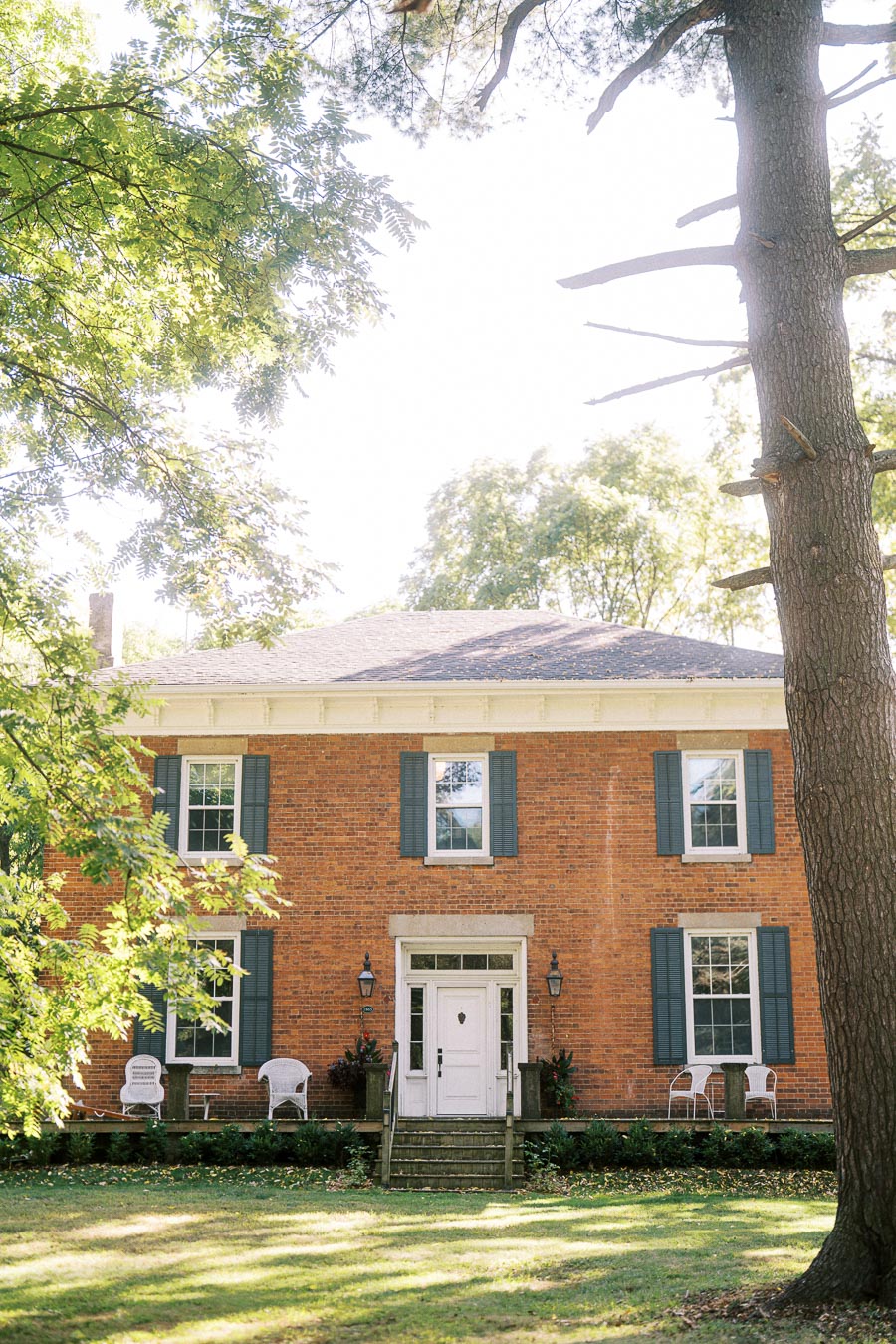 Front view of a charming brick house with large windows and blue shutters, surrounded by lush green trees, featuring a cozy porch with white chairs.