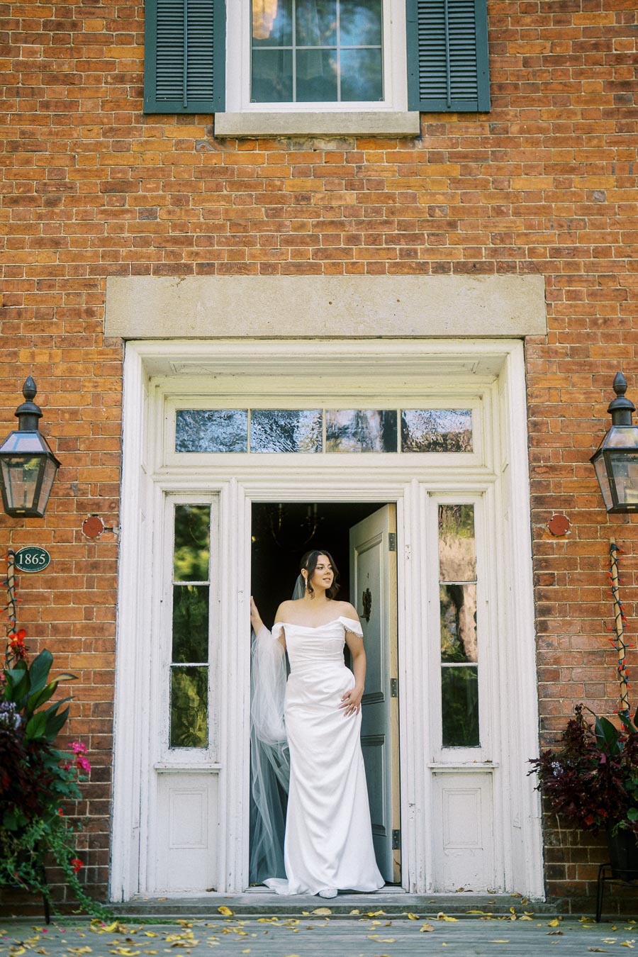 Elegant bride in off-the-shoulder white gown standing in a doorway of a historic brick building with green shutters.