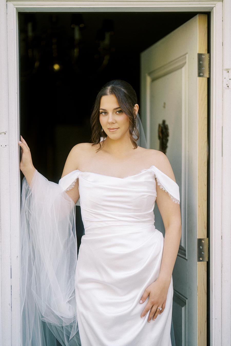 Elegant bride wearing off-shoulder white wedding dress standing in doorway, showcasing wedding fashion and bridal beauty.