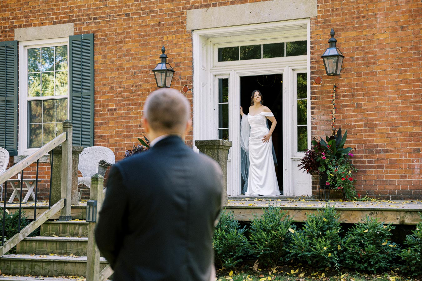 A bride in an elegant white dress stands in a doorway, looking towards a man in a suit with his back turned, outside a charming brick building with green shutters and surrounded by plants.