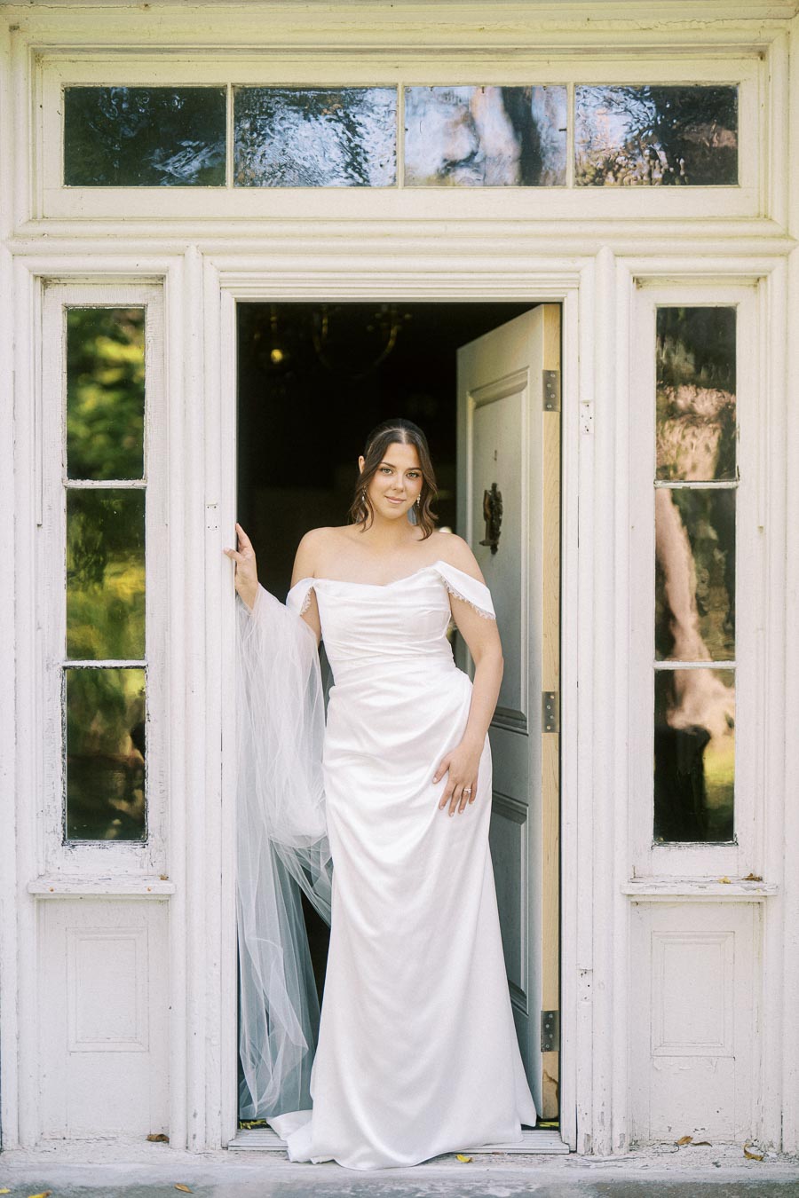 Bride in an elegant white wedding dress standing in a vintage doorway, showcasing off-the-shoulder gown design and flowing veil.
