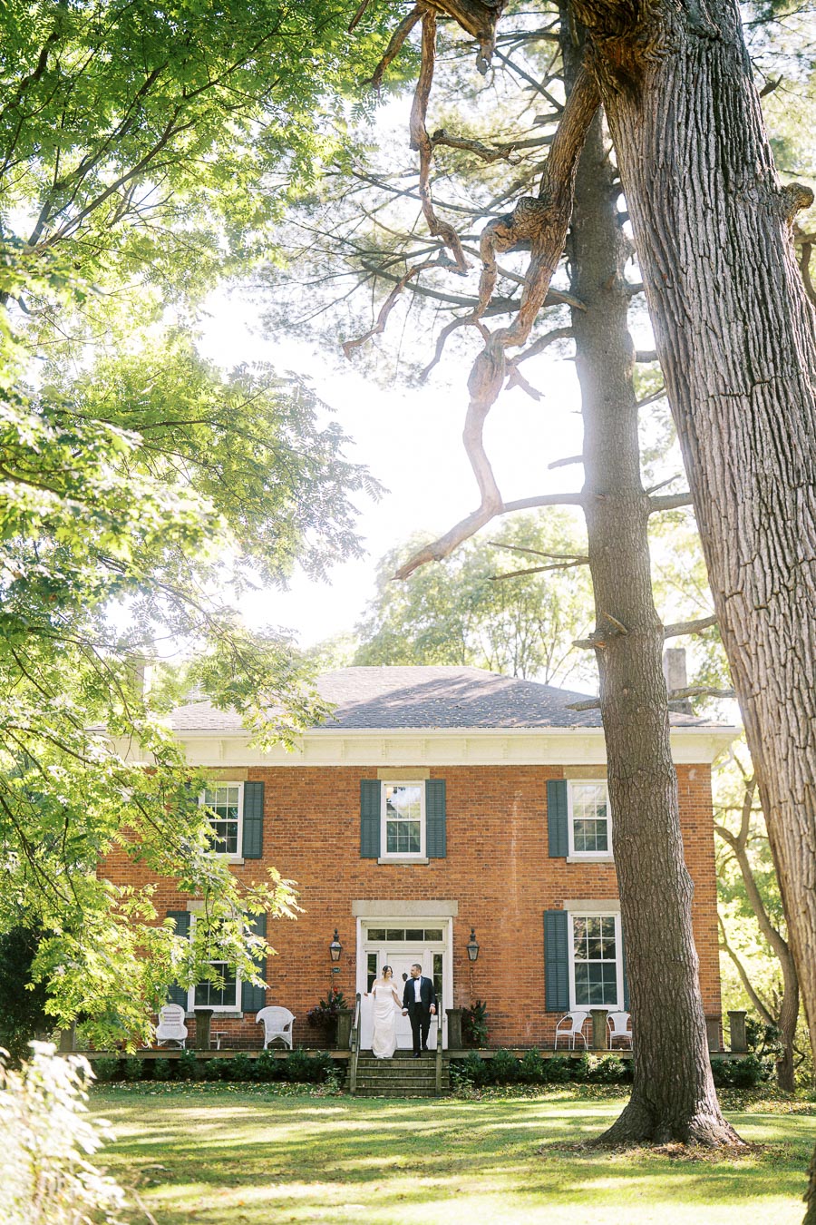 Elegant brick house surrounded by lush greenery with a couple standing on the front steps, exuding charm and tranquility, perfect for weddings or retreats.