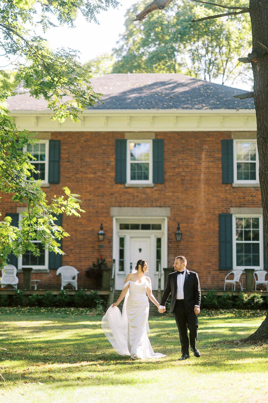Elegant couple in wedding attire walking hand-in-hand on a sunny day in front of a classic brick house with blue shutters.