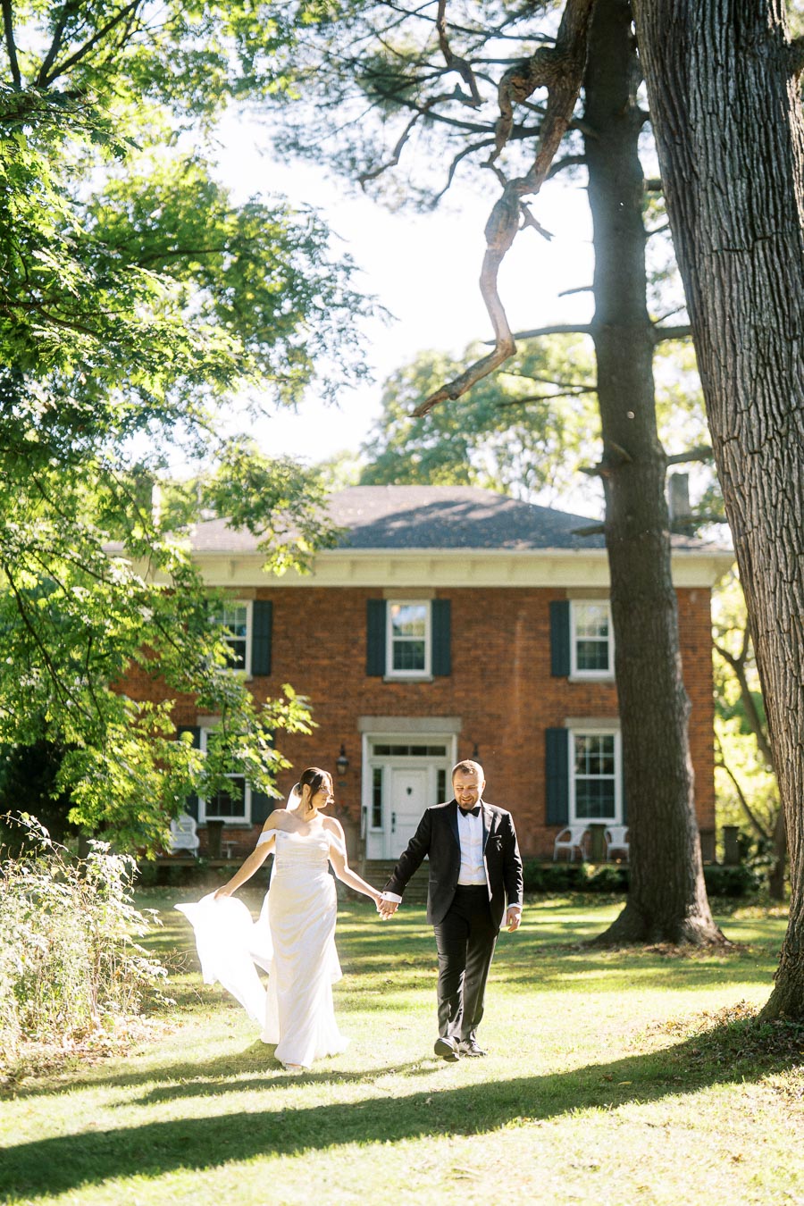 Bride and groom walking hand in hand on a sunny day in front of a historic brick house surrounded by lush greenery.