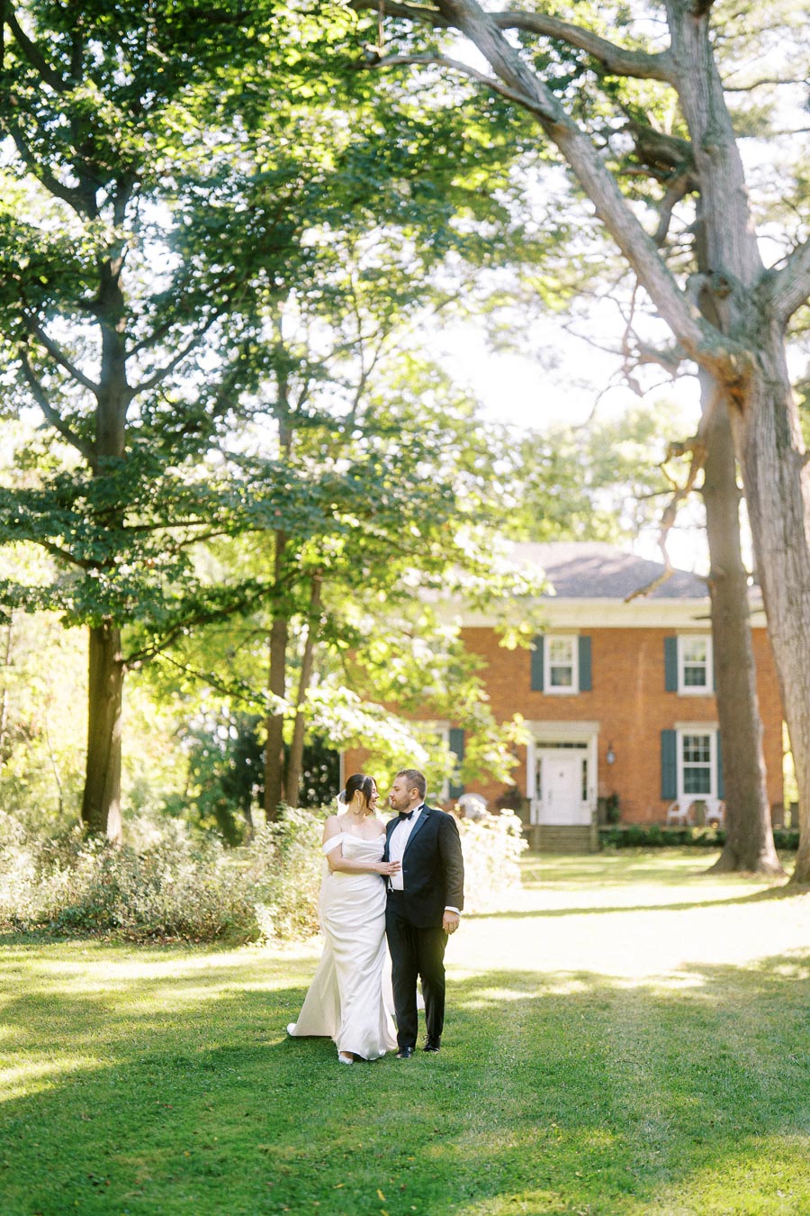 A bride and groom walking together on a lush green lawn, surrounded by tall trees, with a brick house in the background, on a sunny day.