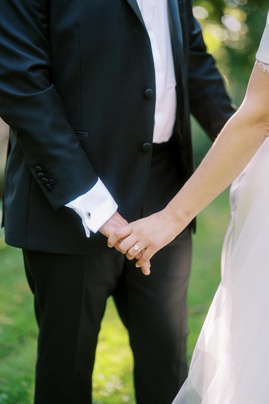 A bride and groom holding hands, showcasing a wedding ring, with the groom in a black suit and the bride in a white dress, standing on a grassy outdoor backdrop.