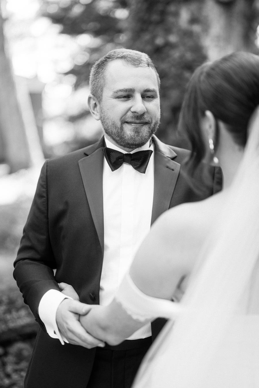 Black and white photo of a groom in a tuxedo smiling at the bride, holding hands during their outdoor wedding ceremony.