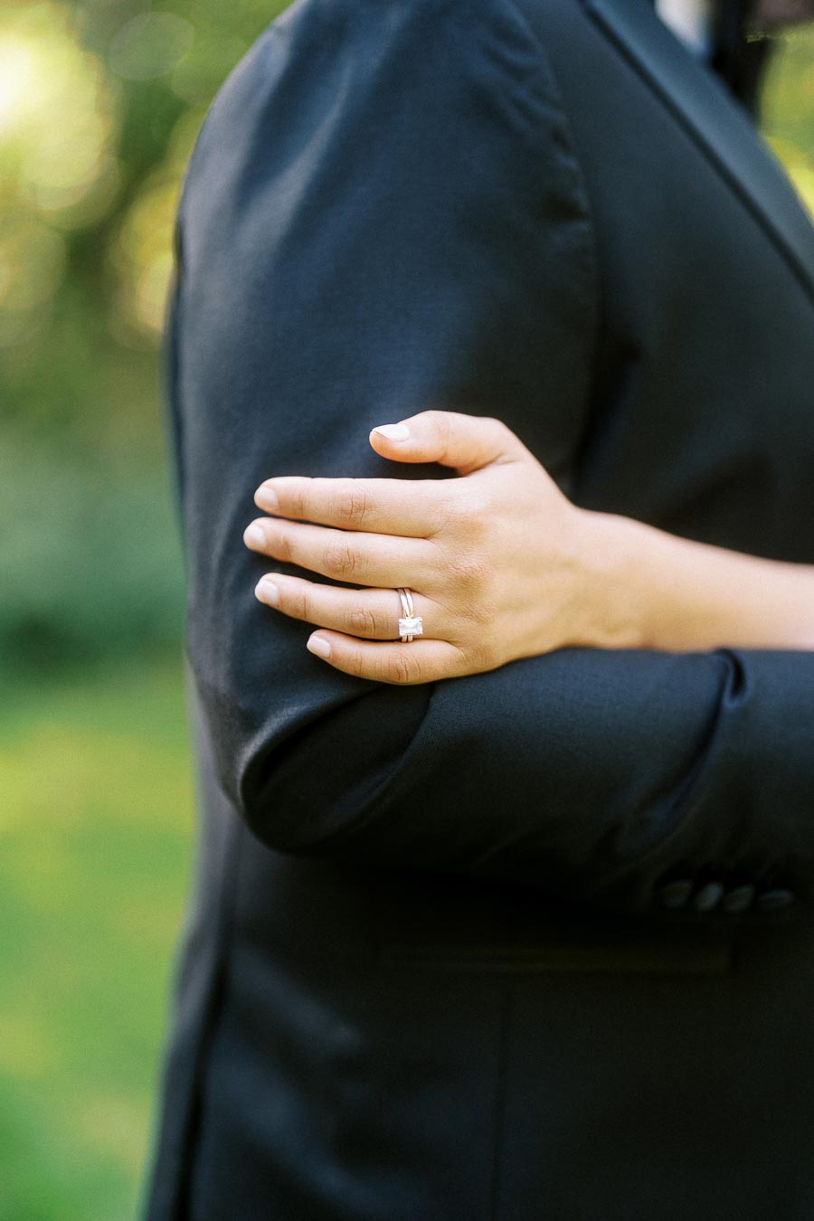 A close-up image of a person wearing a suit with another person’s hand resting on their arm, showcasing an elegant engagement ring, set against a blurred outdoor background with lush greenery.