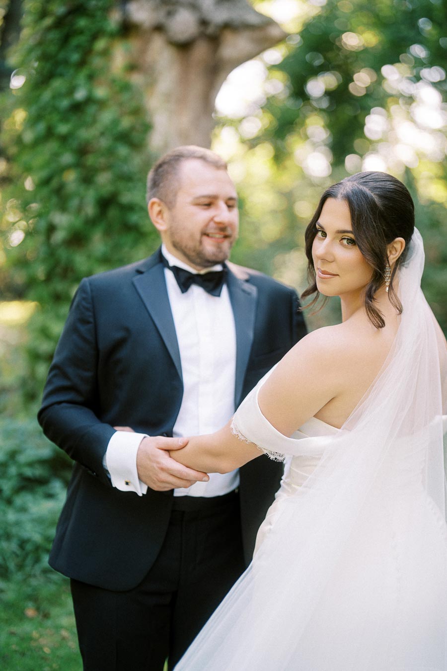 Elegant bride and groom holding hands in a lush green garden, with the bride in a stylish off-shoulder gown and veil, and the groom in a classic black tuxedo, creating a romantic wedding scene.