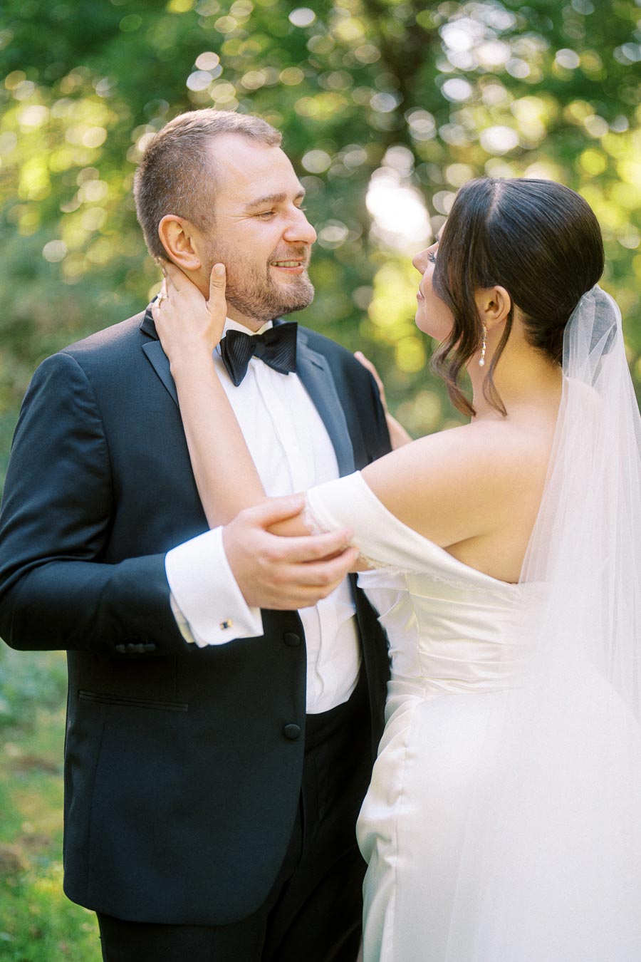 A bride and groom smiling at each other during their outdoor wedding, with the groom in a black tuxedo and the bride in an off-the-shoulder wedding gown, surrounded by a lush green forest background.