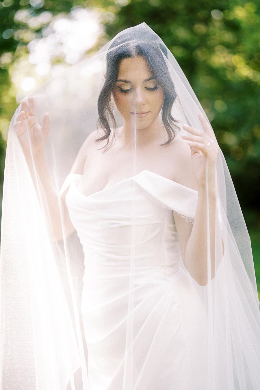 Bride wearing an elegant white wedding dress and veil, standing in a sunlit garden with soft-focus greenery in the background.