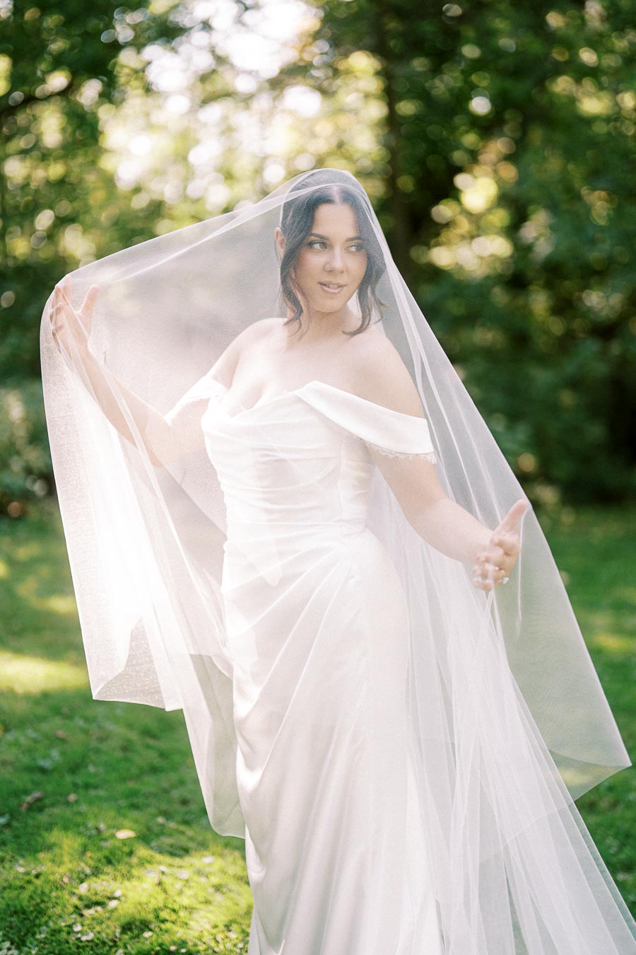 A bride wearing an off-the-shoulder white wedding dress and sheer veil, standing outdoors in a lush garden setting with sunlight filtering through the trees.