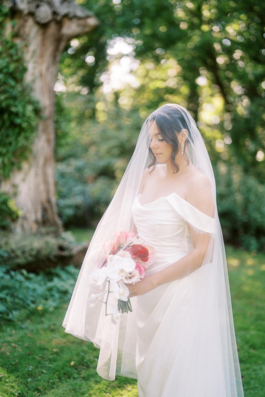 Bridal portrait in a lush garden setting, featuring a bride in an elegant off-shoulder white gown and veil, holding a bouquet of pink and white flowers.