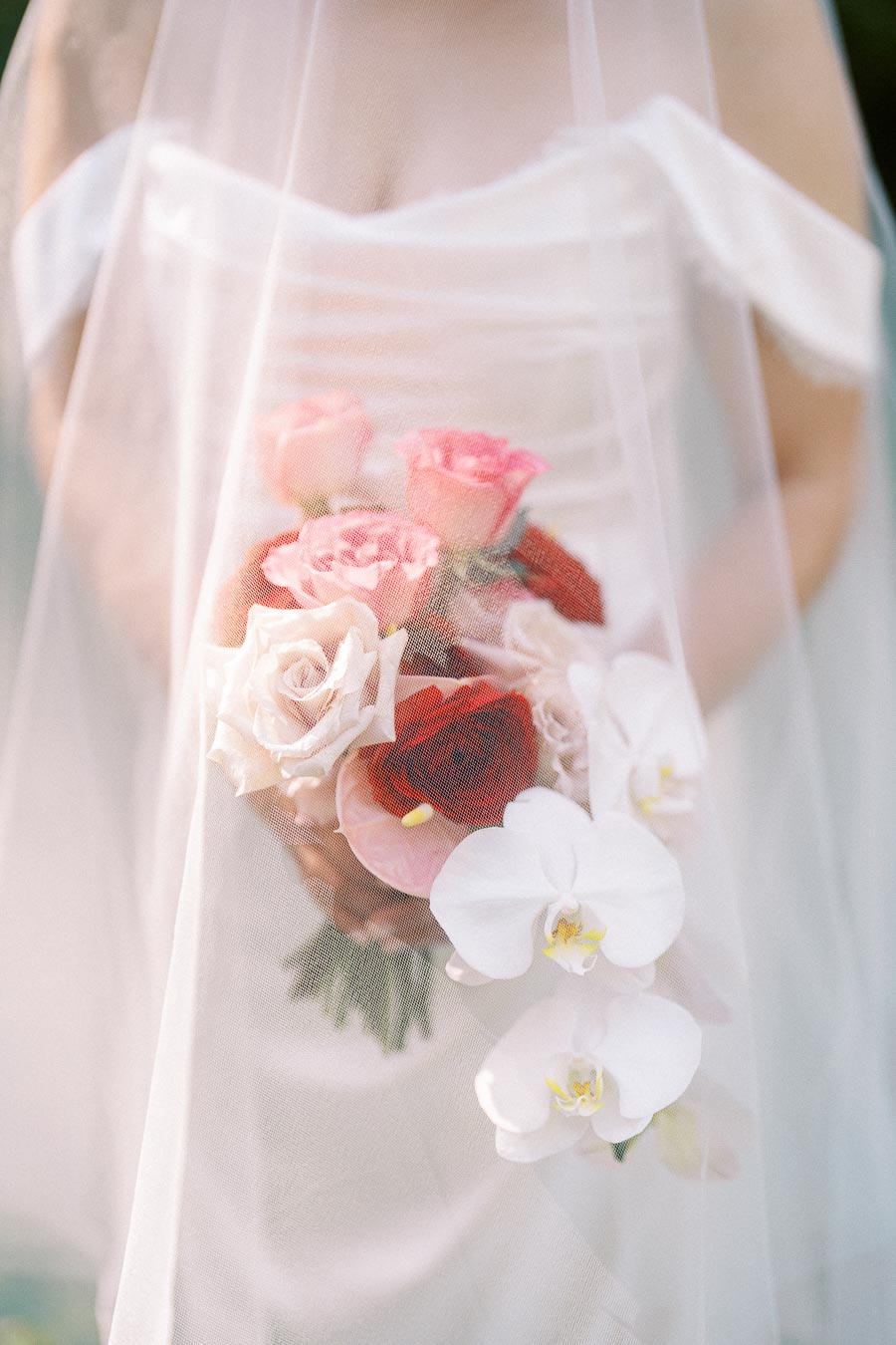A bride holding a bouquet of pink and red roses, white orchids, and other flowers, partially covered by a flowing veil, capturing an elegant and romantic wedding moment.