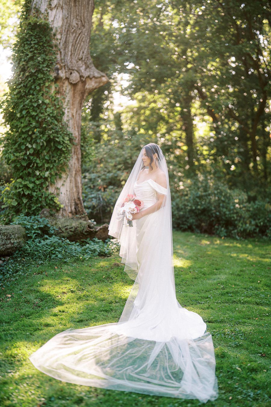 A bride in a flowing white wedding dress and veil stands gracefully on a lush green lawn, holding a bouquet of pink and white flowers, with a large tree and serene garden setting in the background.