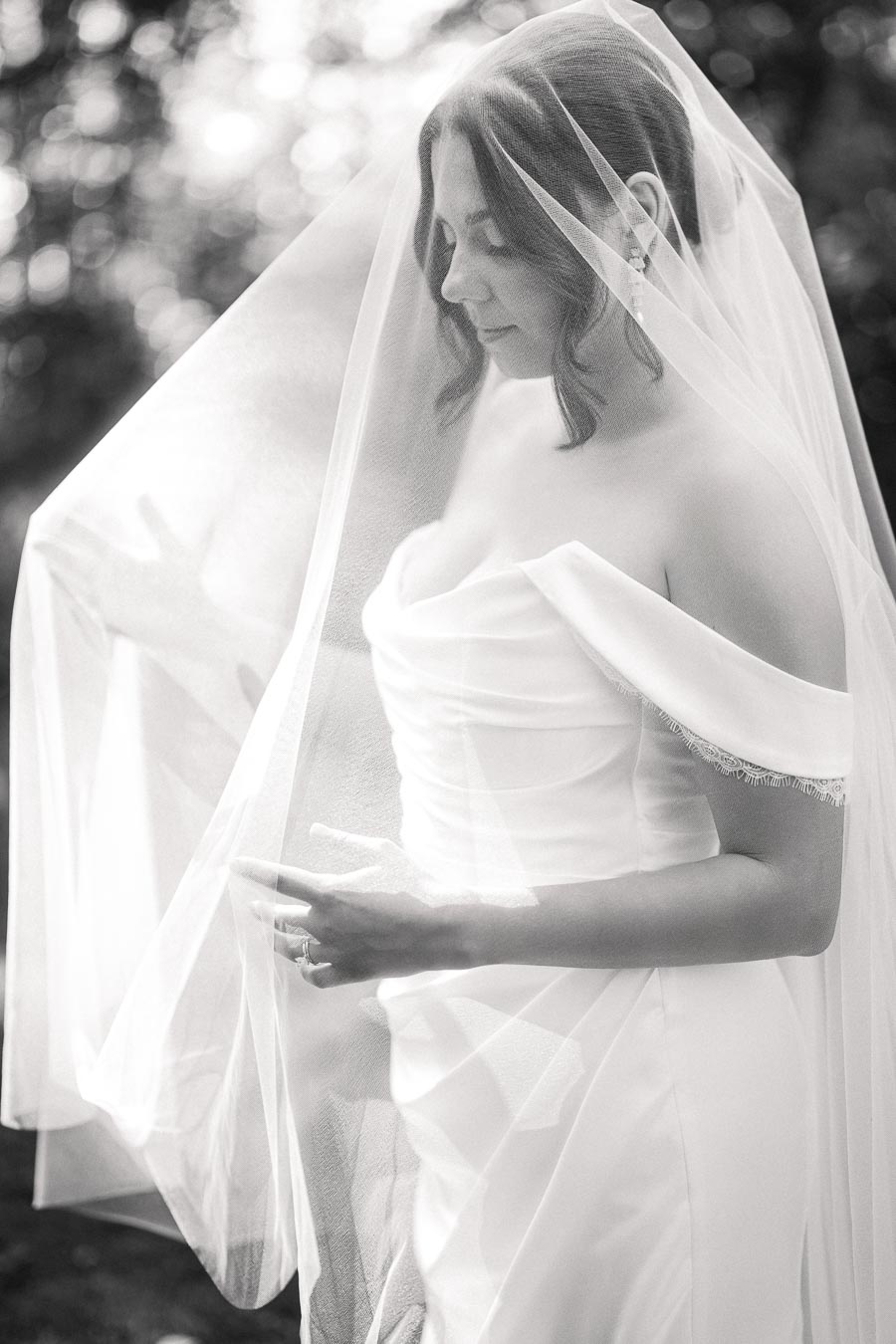 Black and white portrait of a bride in an elegant off-shoulder wedding dress, with a delicate veil covering her face, standing serenely outdoors.