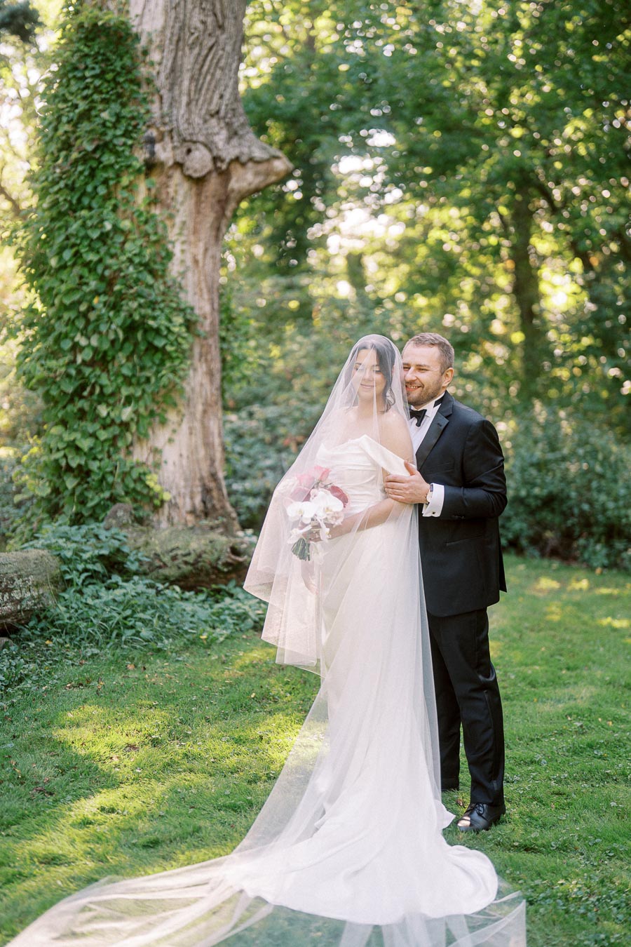 A bride in a flowing white dress and veil holds a bouquet while standing in a lush garden with her groom in a black tuxedo.