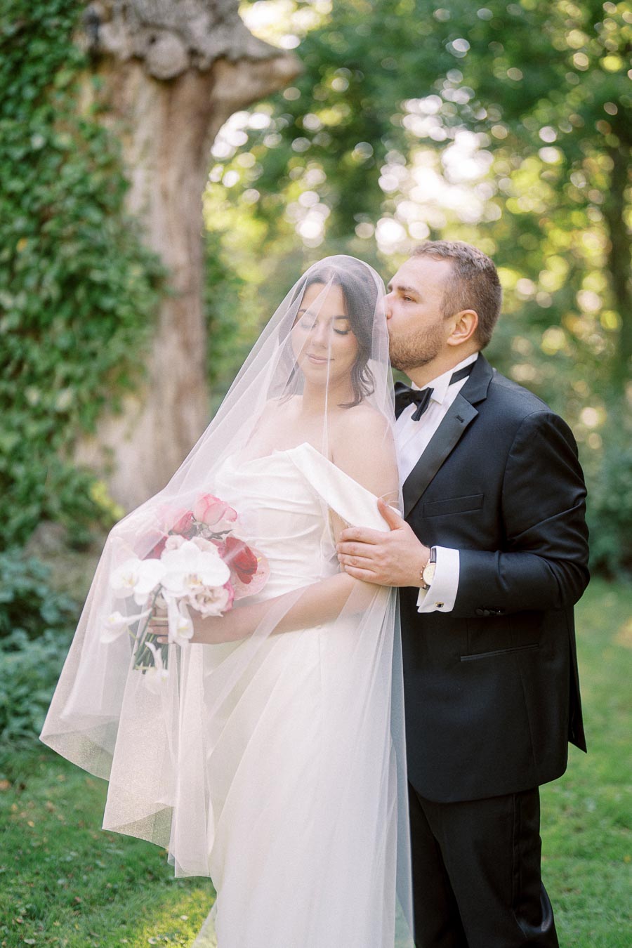 Bride and groom embrace in a romantic outdoor wedding setting, with the groom gently kissing the bride's forehead under her veil, surrounded by lush greenery.