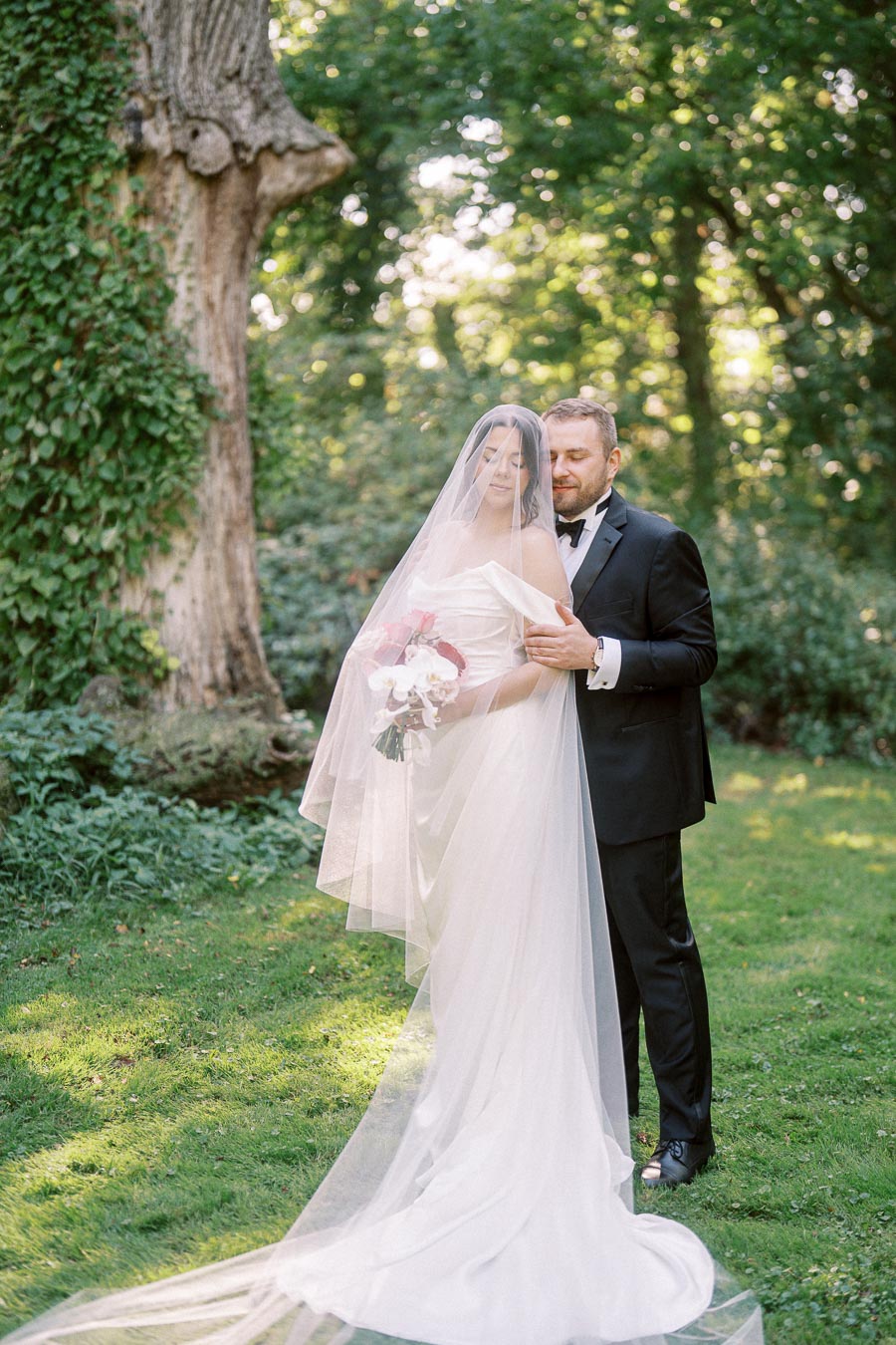 A bride in a flowing white gown and veil holds a bouquet while standing with a groom in a black tuxedo in a lush green garden, under a large tree, capturing a romantic wedding moment.