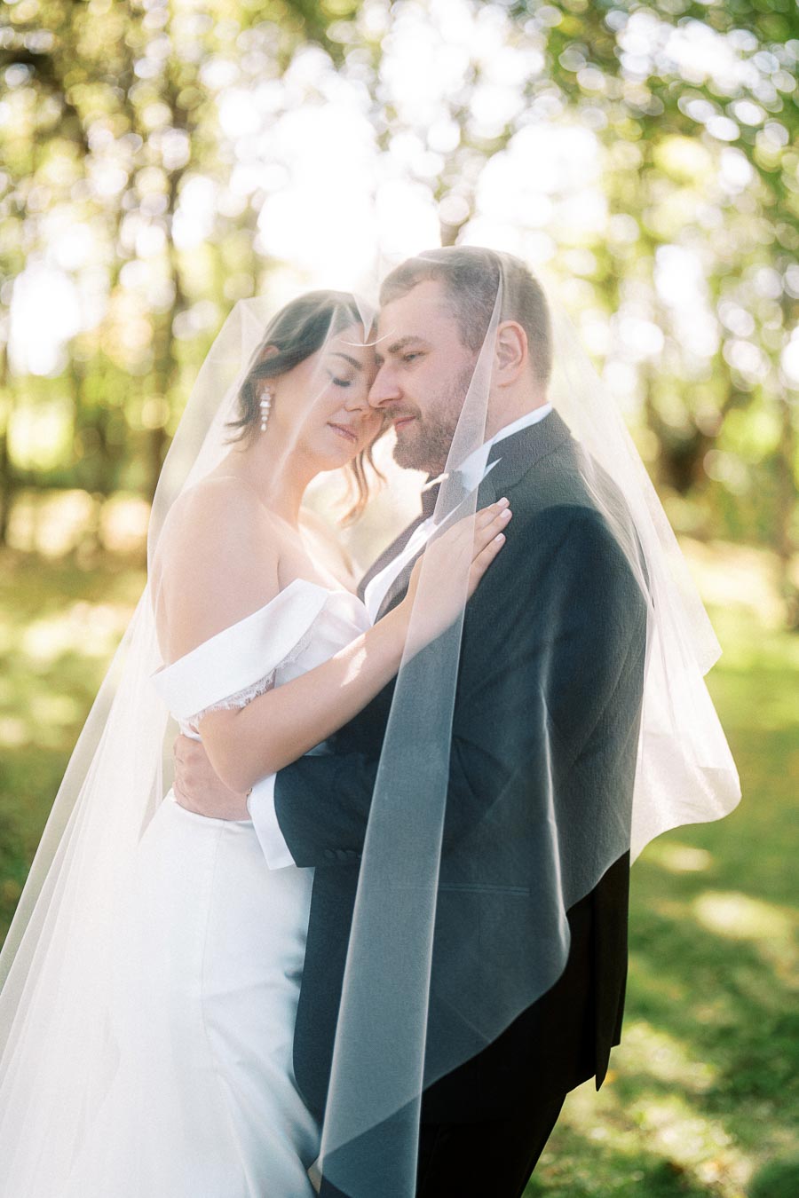 A bride and groom embracing under a veil in a sunlit garden, capturing a romantic wedding moment.