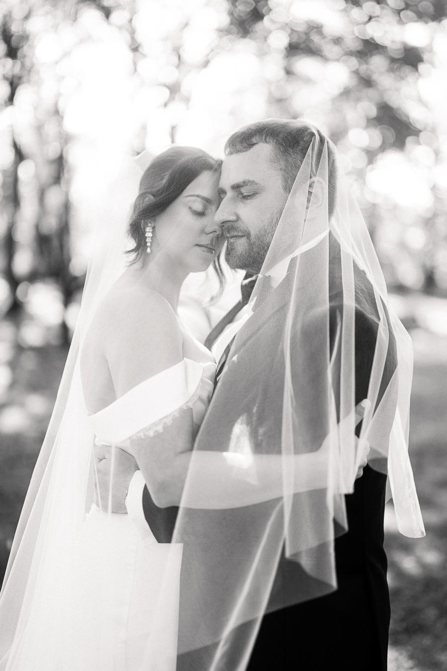 A black and white photograph of a bride and groom embracing tenderly under a veil in an outdoor setting, conveying romance and intimacy.