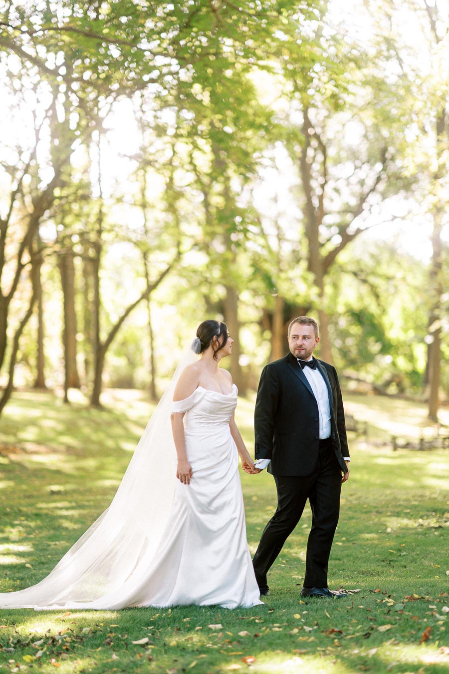 A bride and groom holding hands while walking through a sunlit forested park, with the bride wearing an elegant white gown and the groom in a black tuxedo.