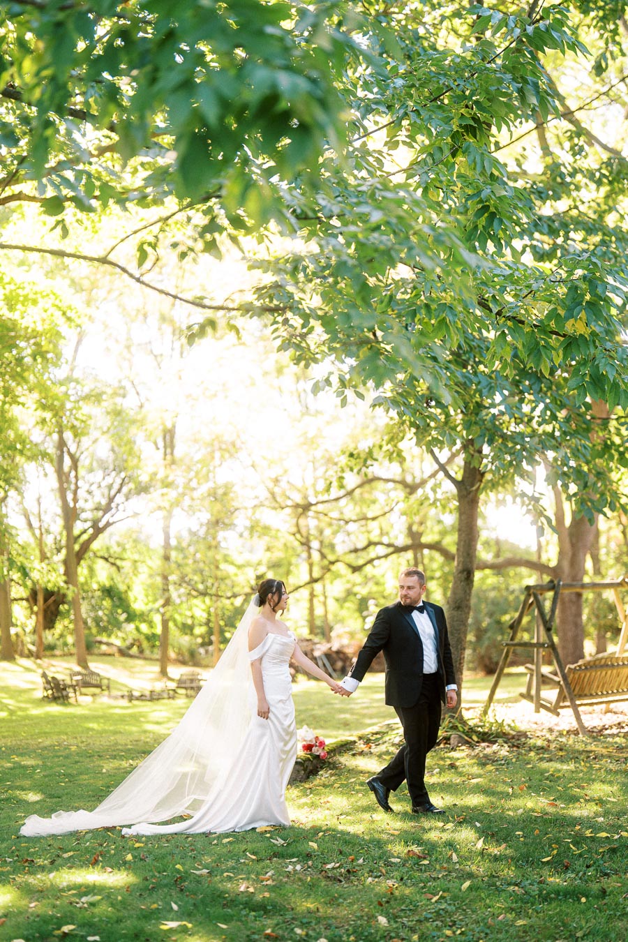 A bride in a white gown with a flowing veil holds hands with a groom in a black suit as they walk through a sunlit, lush green garden, surrounded by tall trees and a wooden swing.