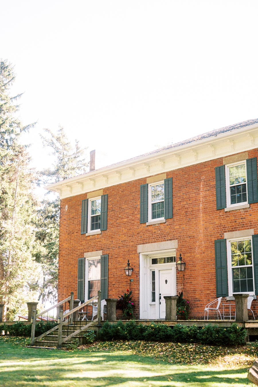 Historic brick house with white trim and green shutters surrounded by lush greenery, featuring a quaint front porch and classic architecture.