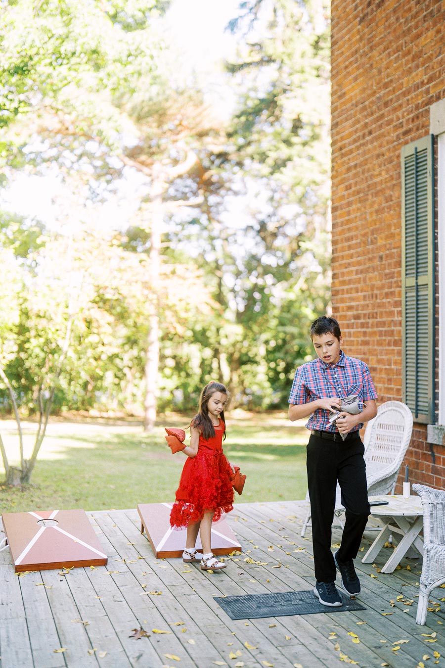 Children playing cornhole on a sunny wooden deck, with a vibrant green garden in the background.
