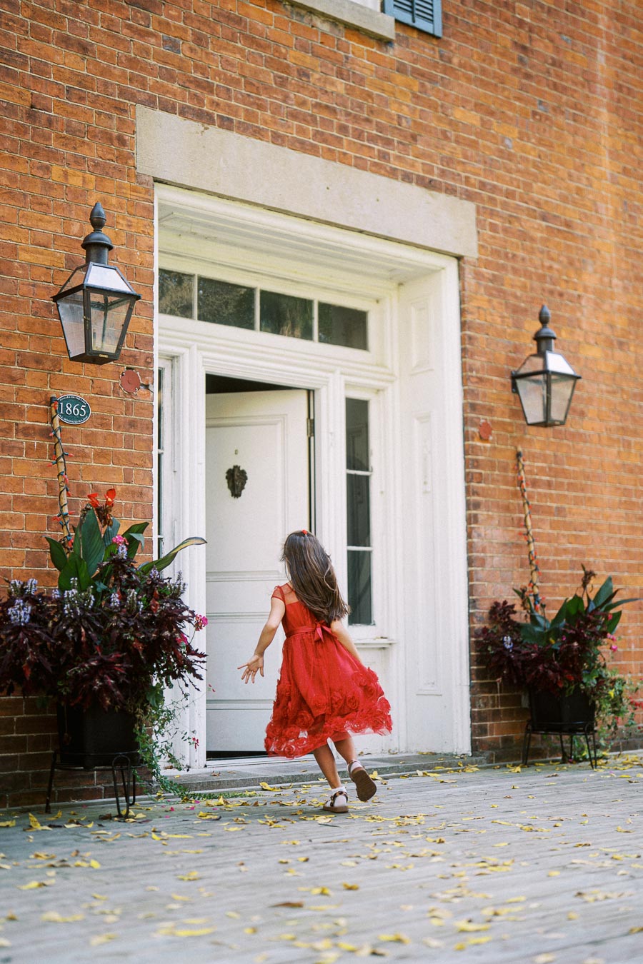 A young girl wearing a red dress joyfully runs towards the entrance of a historic brick building, adorned with green plants and vintage lanterns on either side of the door.