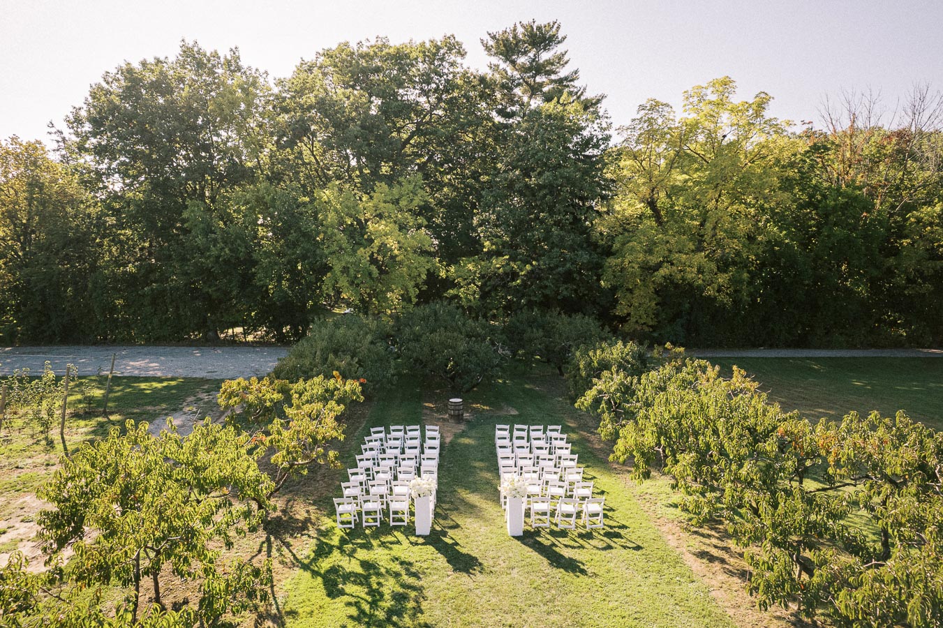 Aerial view of an outdoor wedding ceremony setup with white chairs arranged in rows on a lush green lawn, surrounded by trees under clear blue skies.