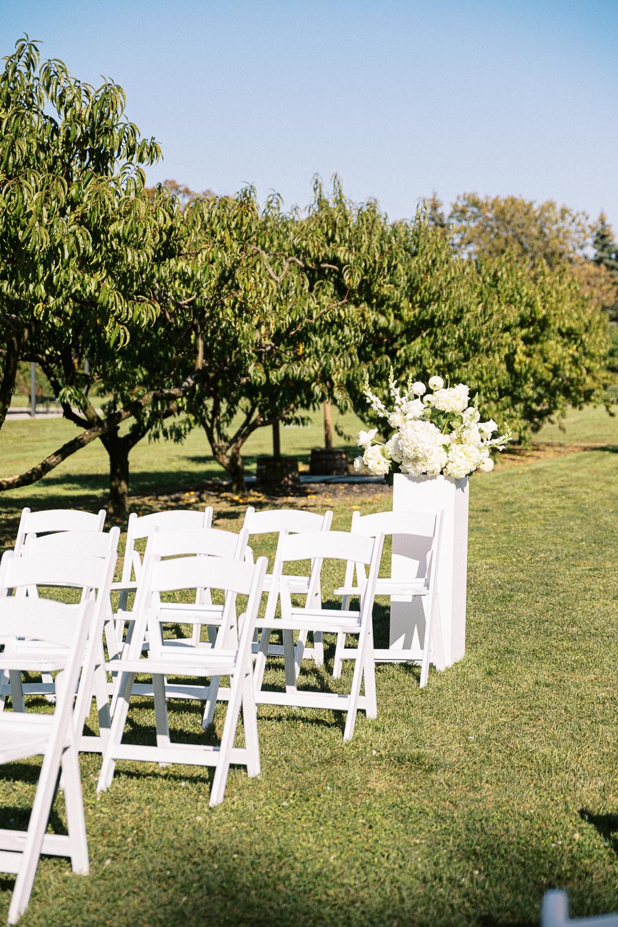 Outdoor wedding ceremony setup with white folding chairs arranged in rows on green grass, next to a floral arrangement on a white pedestal under sunny skies.