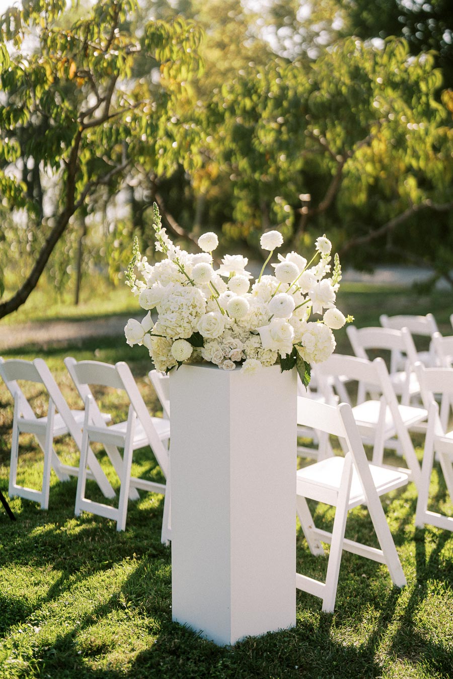 White floral arrangement on a tall pedestal with white folding chairs arranged outdoors for a wedding ceremony, surrounded by lush green trees and grass.