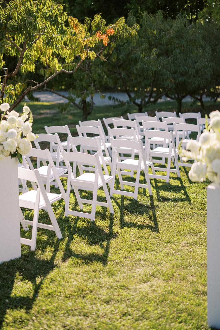 Outdoor wedding ceremony setup with rows of white folding chairs on a lush green lawn, surrounded by trees and decorated with white floral arrangements.