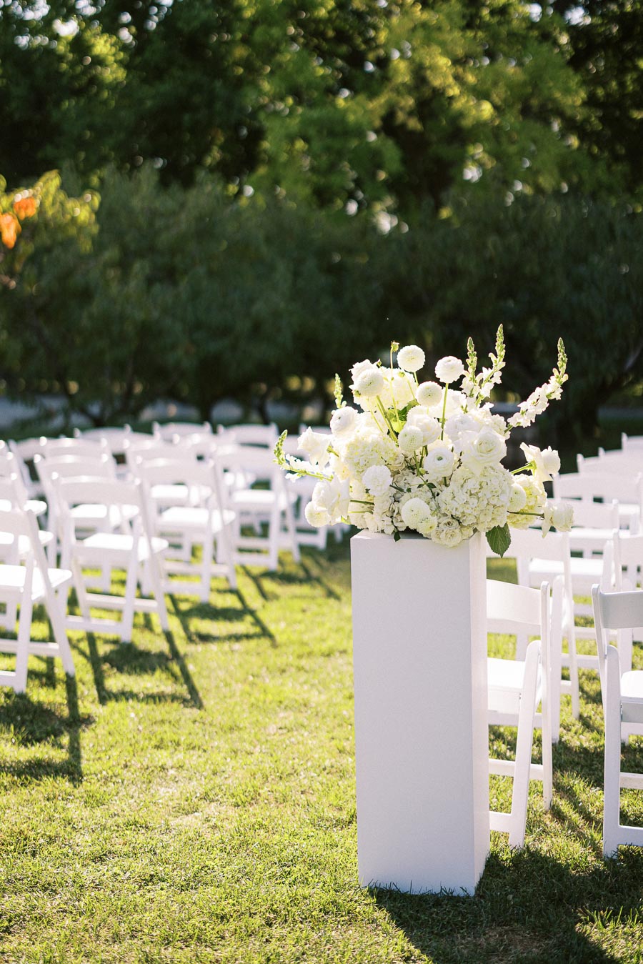 Elegant outdoor wedding ceremony setup with white chairs and a tall floral arrangement featuring white flowers on a grassy lawn.
